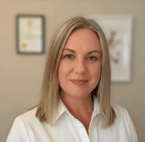 Professional headshot of a woman with straight blonde hair and blue eyes, wearing a white blouse, in an office with framed certificates on the wall.