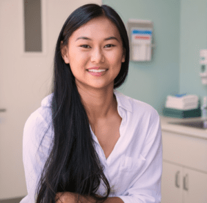 Smiling young woman with long black hair in a white button-down shirt, in a clinical exam room with cabinets in the background.