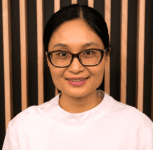 Portrait of a woman with dark hair and burgundy glasses smiling in front of a wooden slat background.]},{