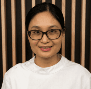 Smiling woman with burgundy glasses and white top, standing against vertical wooden slats backdrop